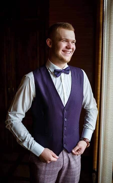 Smiling young man in formal attire for a wedding. Purple shades, vest bowtie and formal shirt